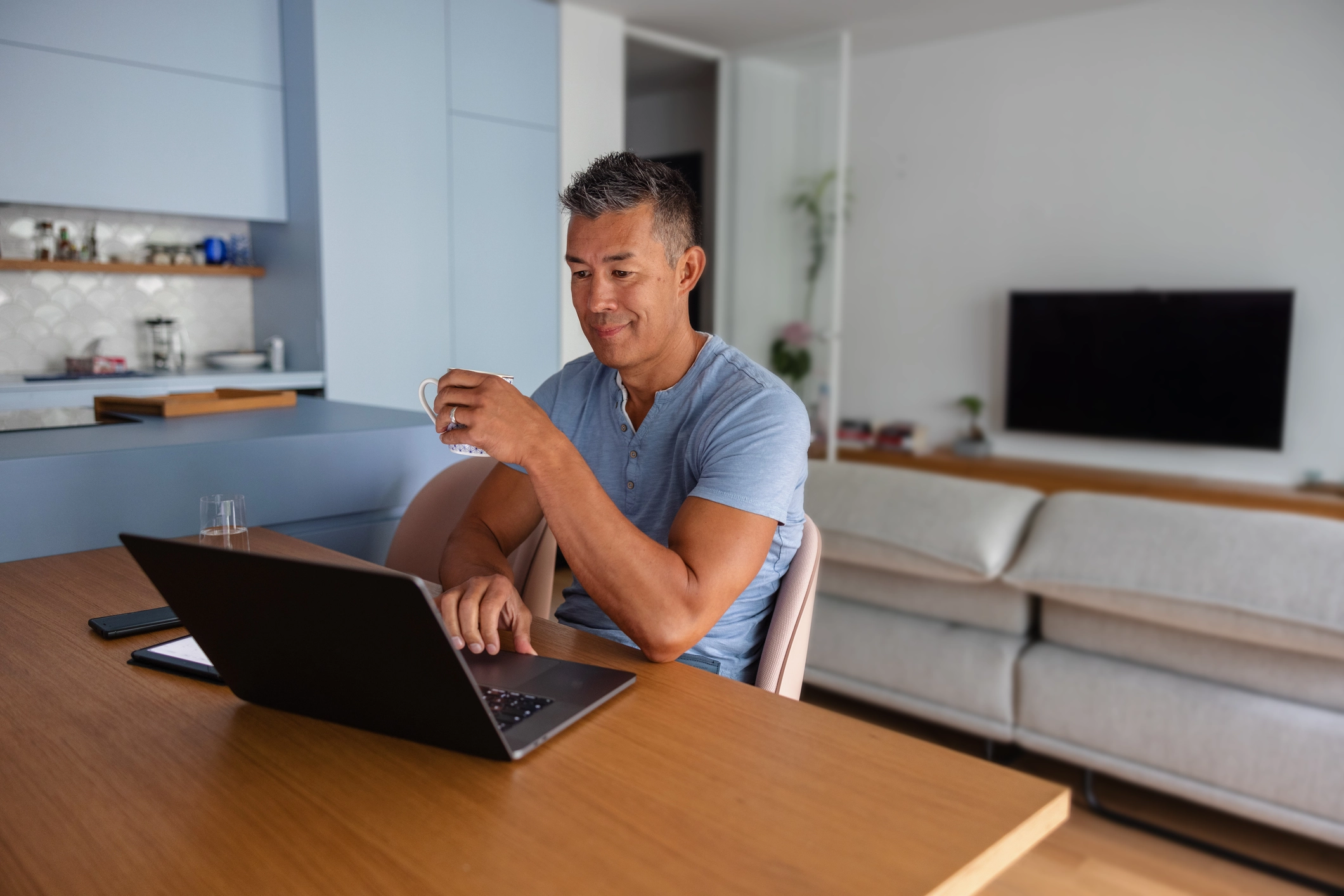 A man smiling while drinking coffee and using a laptop at home